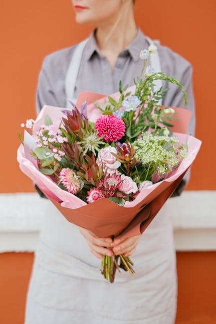 A person dressed in a light grey shirt and white apron holding a large, vibrant floral bouquet wrapped in pink and coral-colored paper. The bouquet features an assortment of fresh flowers including pi