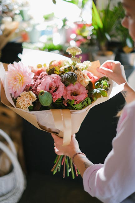 A person holding a large bouquet of fresh flowers, featuring pink peonies, white roses, and purple carnations, wrapped in cream-colored paper with a satin ribbon. The bouquet is arranged with green fo